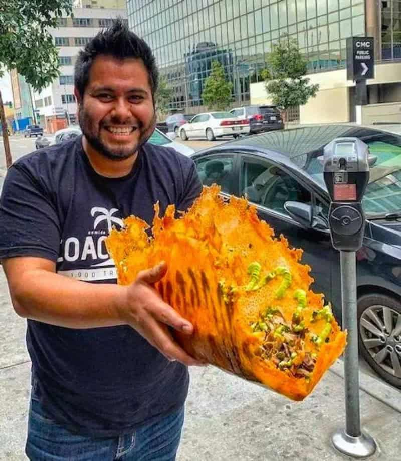 Man smiling holding a large, fiery orange and yellow carved pumpkin on city street.