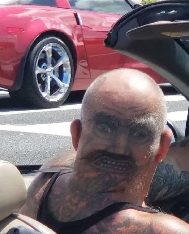 Man with a tattooed face and head, sitting in a convertible car near a red vehicle.