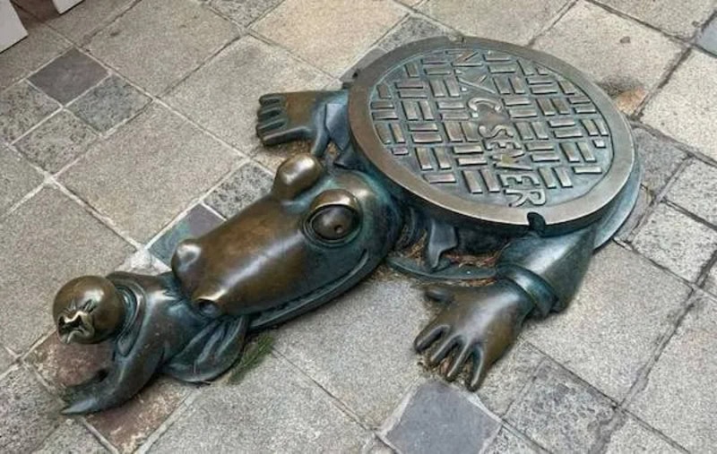 Manhole cover designed as a frog with a round, textured top and detailed limbs, lying on a paved sur.