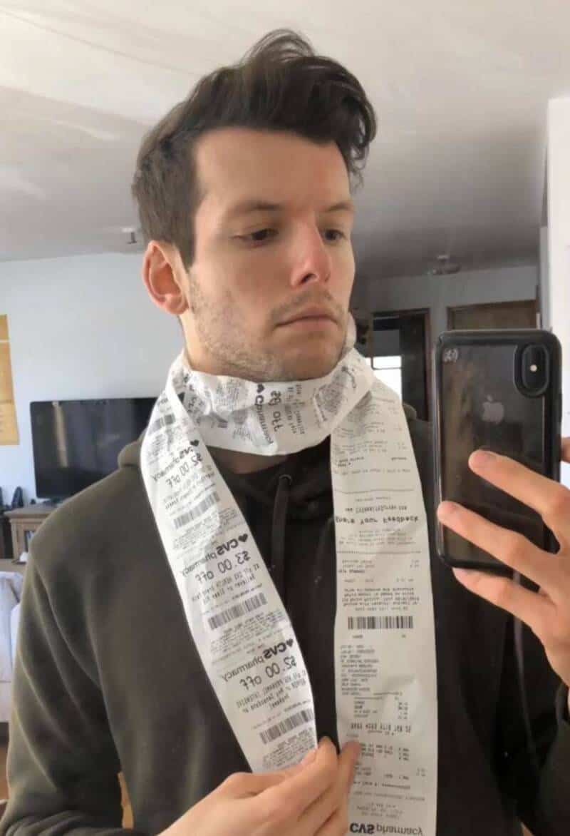 A young man taking a mirror selfie with a smartphone, wearing a scarf made of receipts.