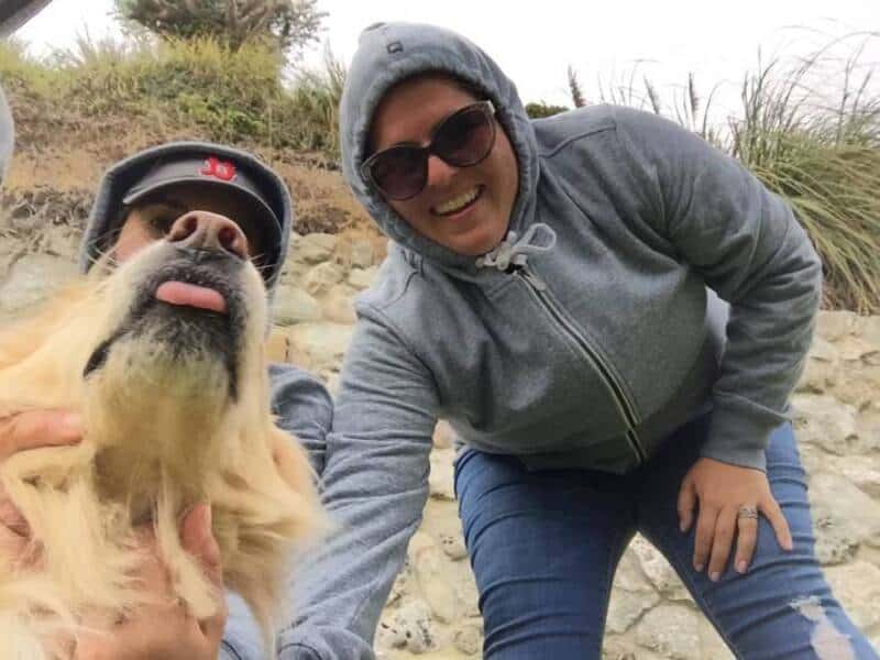 Happy woman in hoodie and sunglasses with golden retriever outdoors on rocky terrain.