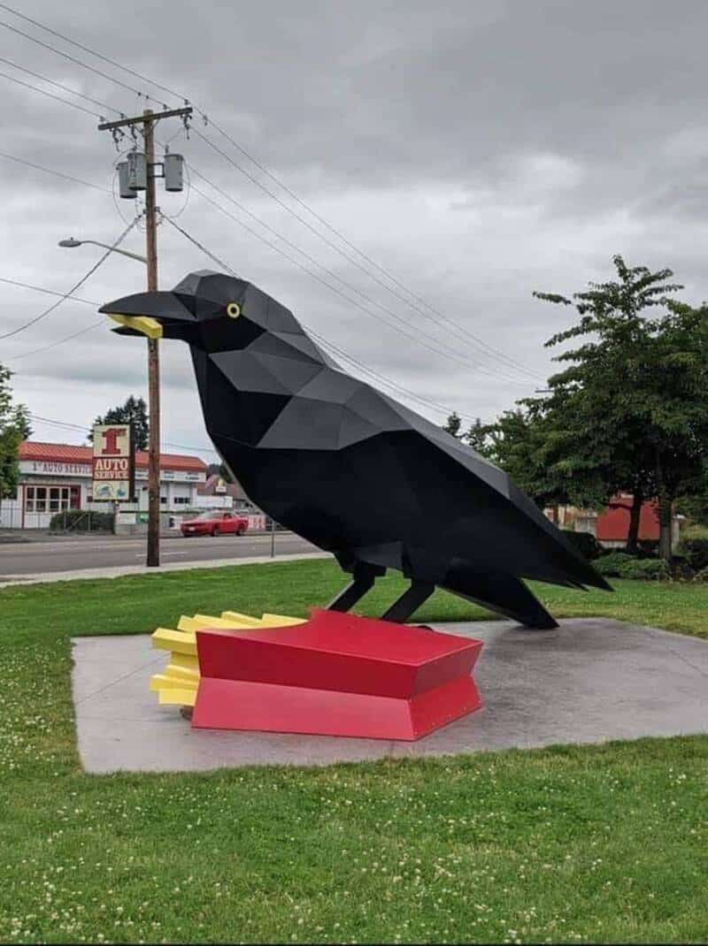 Black crow sculpture with yellow eyes, perched on a red and yellow base, outdoors on a cloudy day.