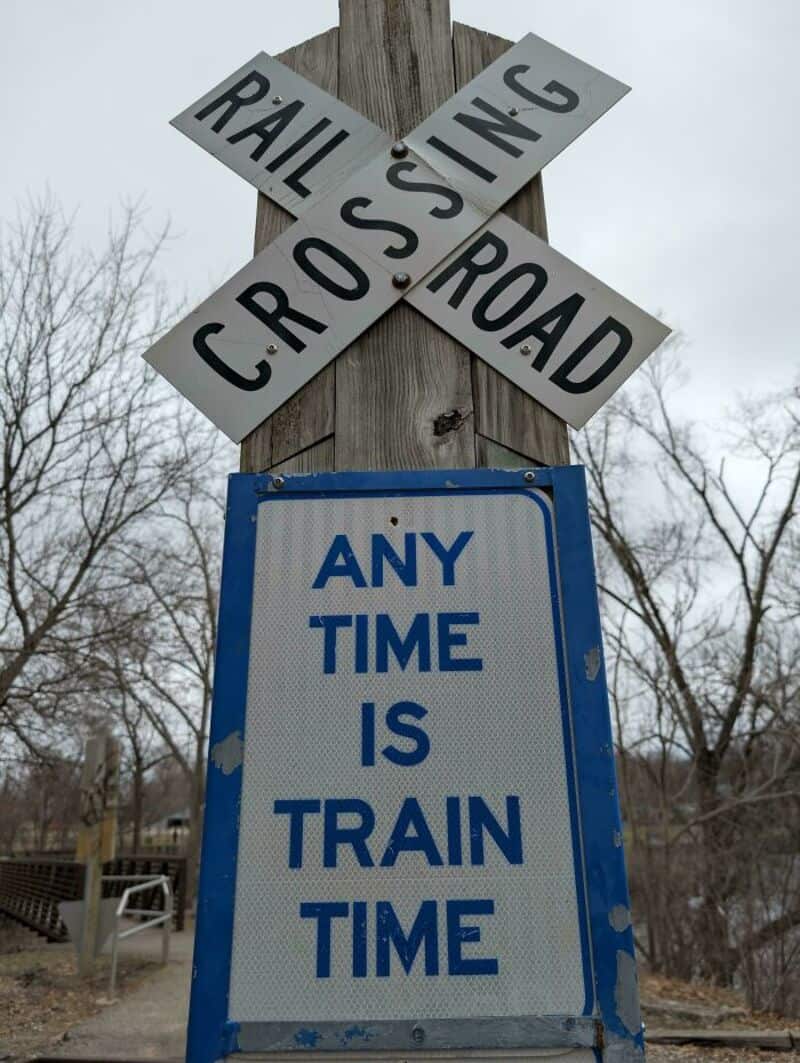 RAIL CROSSING and TRAIN TIME sign on wooden post with trees in background.