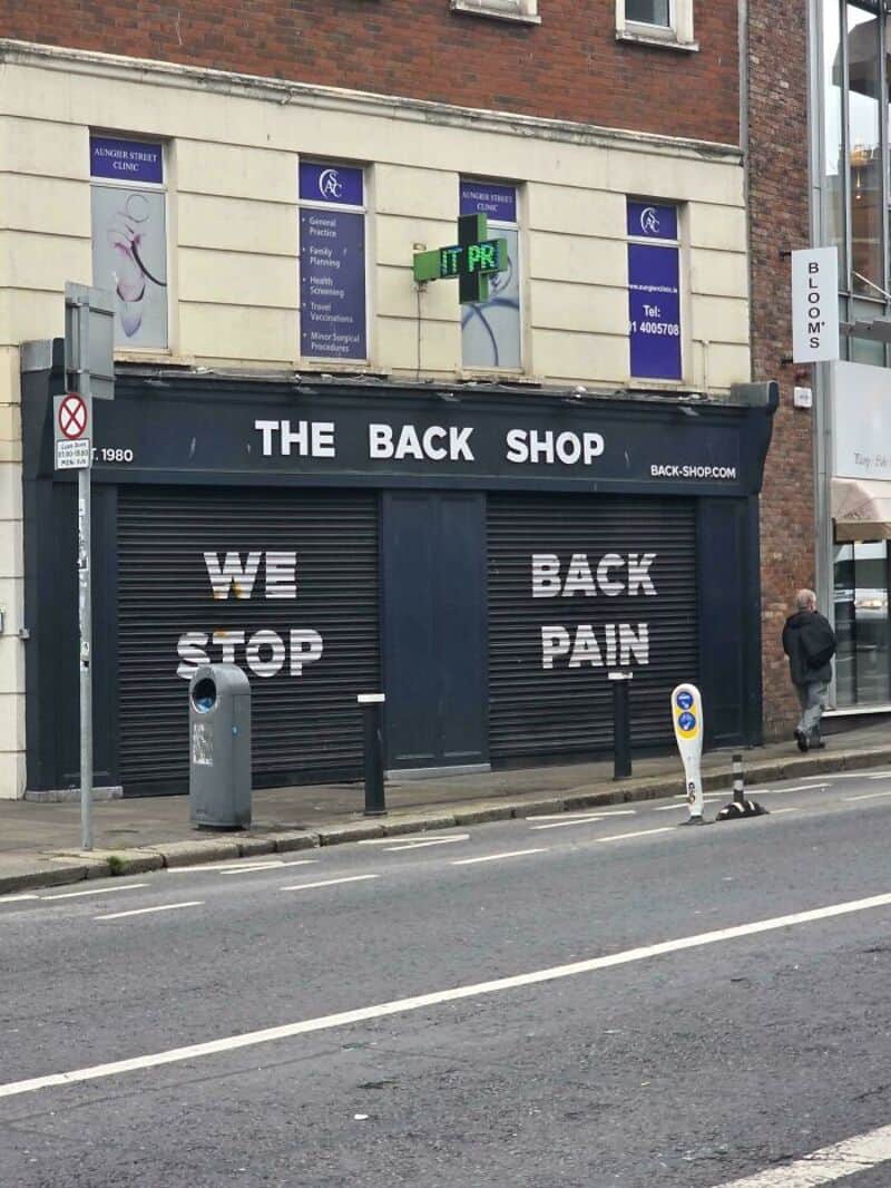 Storefront with a sign reading 'THE BACK SHOP' and graffiti 'WE STOP BACK PAIN' on the shutter.