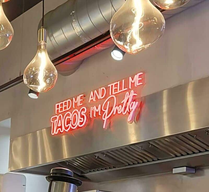Neon sign says 'Home and tell me tacos I'm dirty' in a restaurant ceiling.