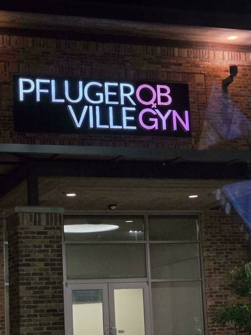 Sign for Pfluger QB Ville GYN clinic on brick wall at night.