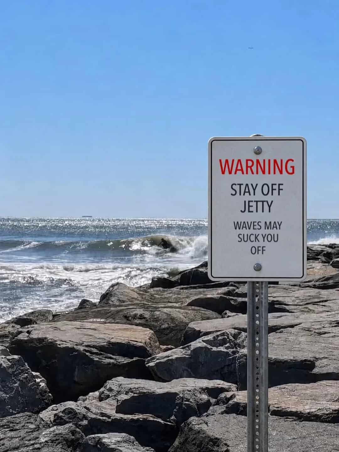 Warning sign on rocks at the beach says 'Stay off jetty. Waves may suck you off.'.