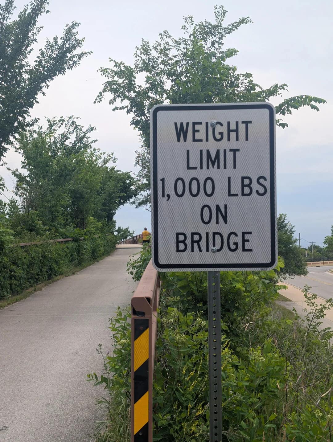 Weight limit 1,000 lbs on bridge sign with trees and road in background.