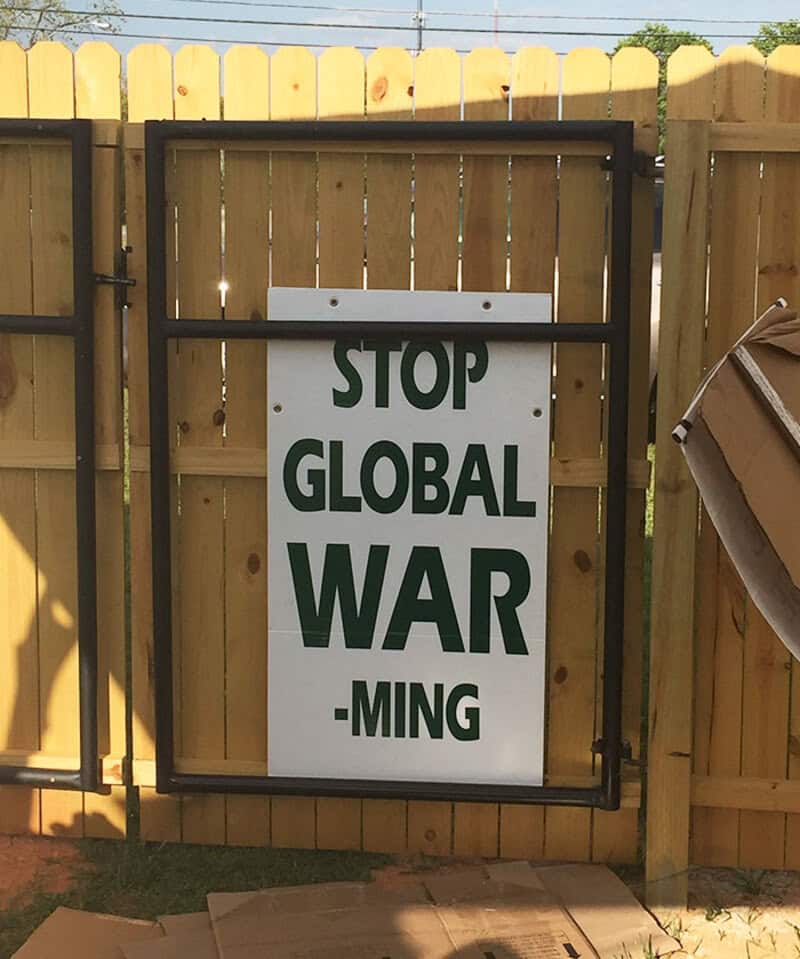 Sign on gate says 'STOP GLOBAL WAR - MING' with a fence and cardboard box nearby.