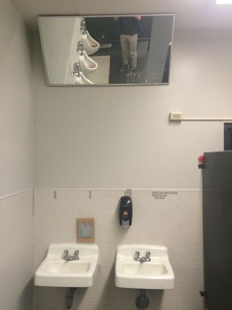 Mirror shows person taking photo, bathroom with two sinks, soap dispenser, and a small picture.