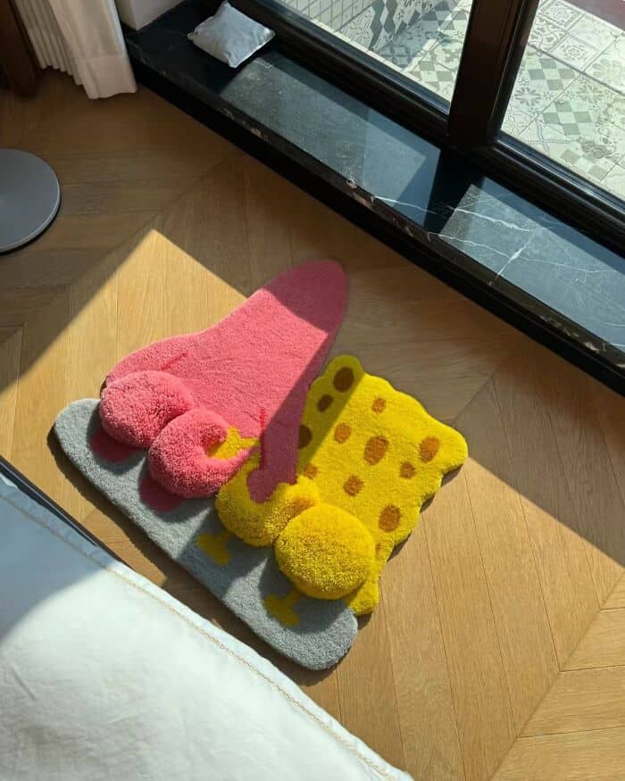 Pink flamingo and yellow sponge cake-shaped slippers on a gray mat near a glass door.