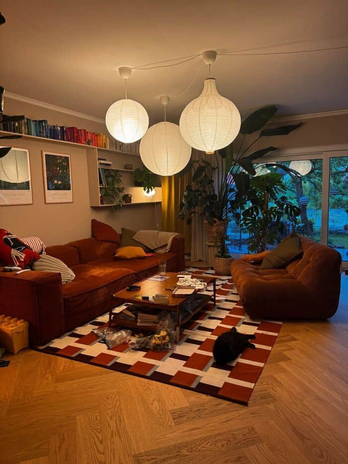 Cozy living room with hanging paper lanterns, bookshelves, and a large window with plants.