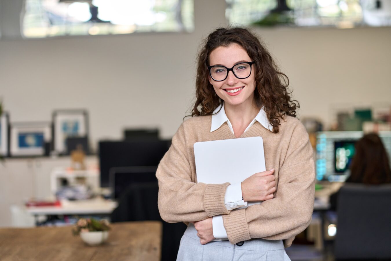 Smiling woman with glasses holding a laptop in an office setting.