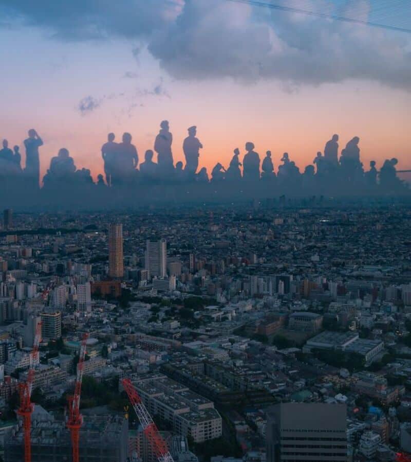 Silhouettes of people standing on a city rooftop at sunset with clouds above.