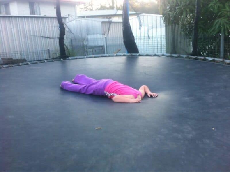 Child lying face down on trampoline in backyard, wearing pink top and purple pants.