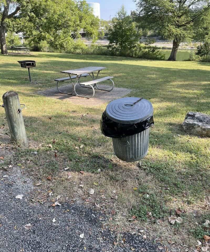 Park scene with picnic table, trash can, and trees in the background.