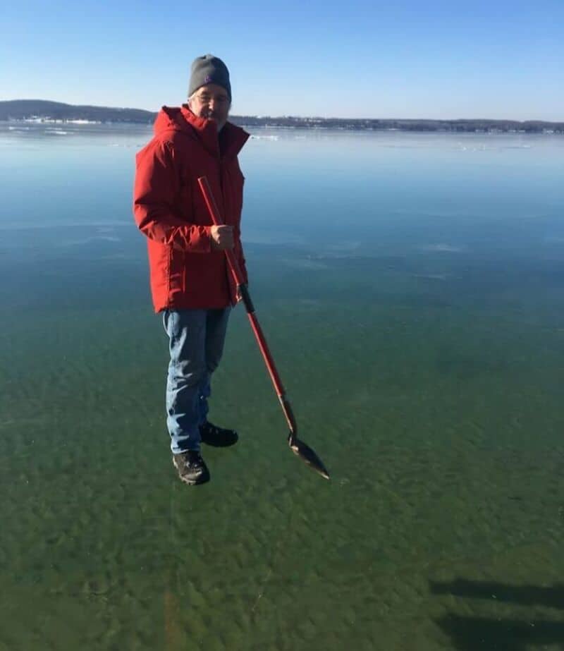 Person in red jacket standing on ice with a shovel, lake and distant shoreline in background.