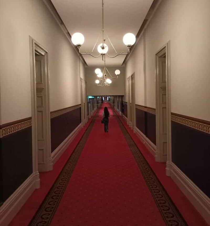 Long corridor with white walls, red carpet, and hanging lights, person walking away.
