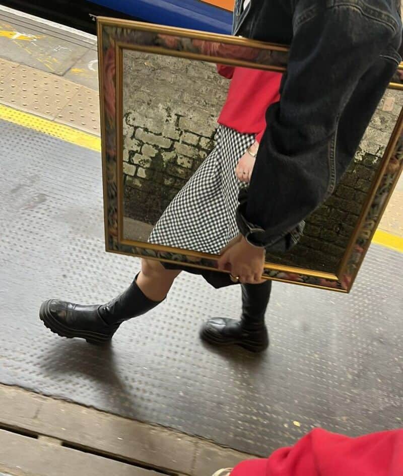 Person holding a mirror reflecting their outfit, including a red top, checkered skirt, and black leg.