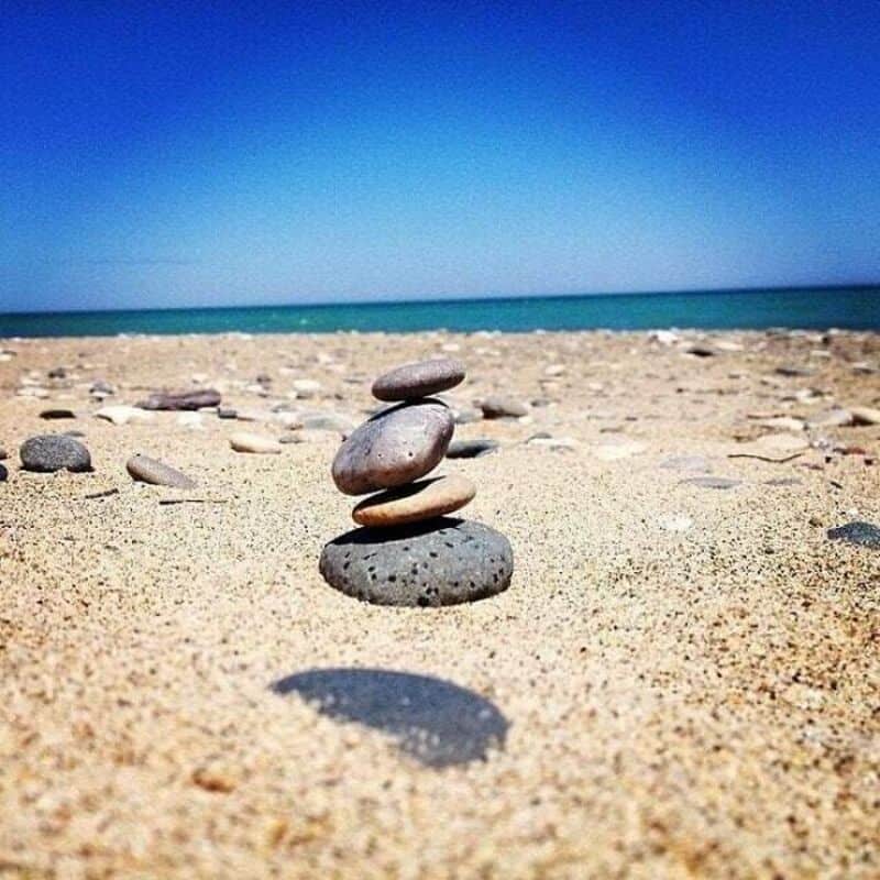 Stacked stones on a sandy beach with the ocean and blue sky in the background.