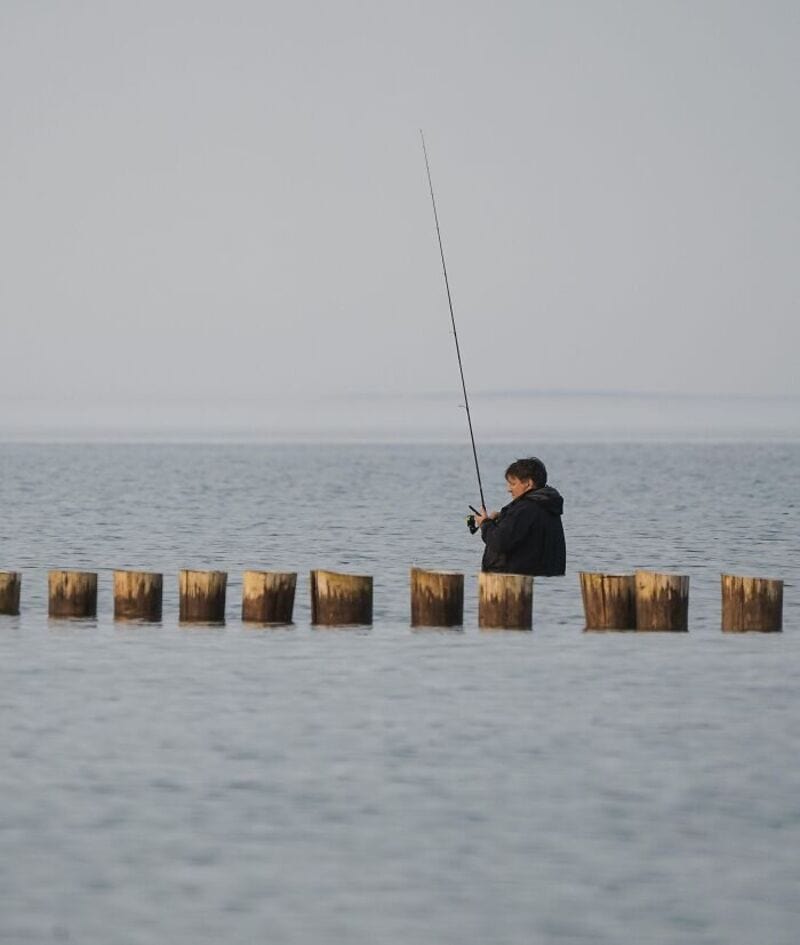 Person fishing on wooden posts in water, overcast sky, calm sea.