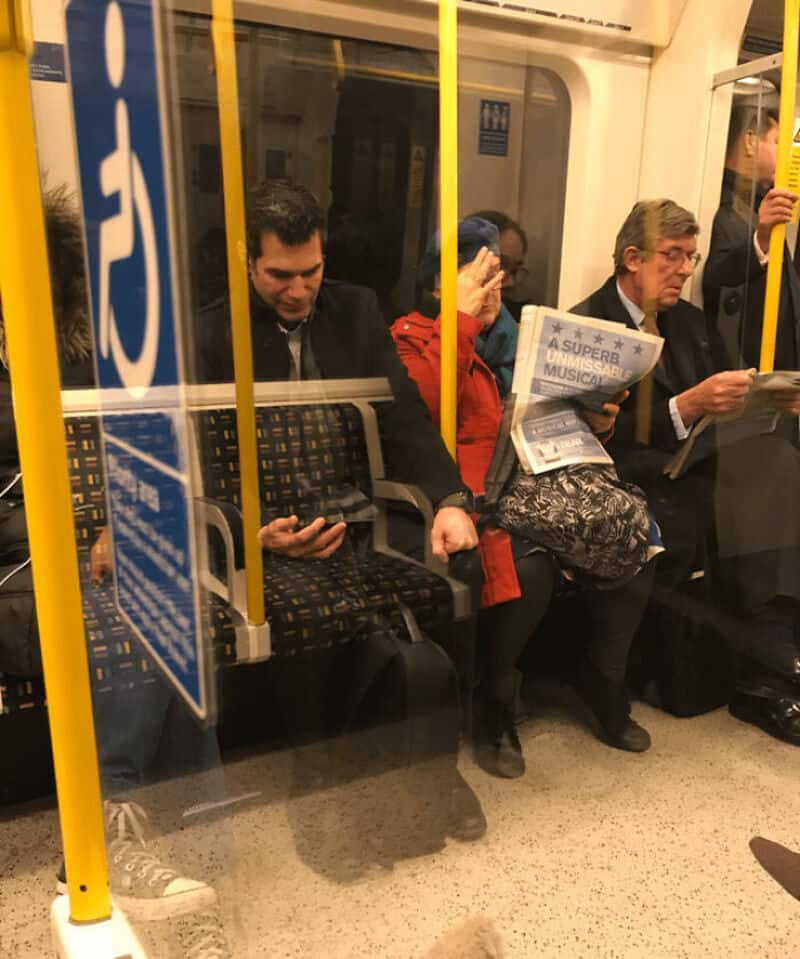 Three people sitting on a train, one reading a newspaper, one looking at a phone, and one with hand.