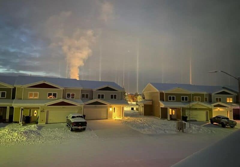 Snowy neighborhood with rainbow and cloudy sky, smoke rising from distant factory.