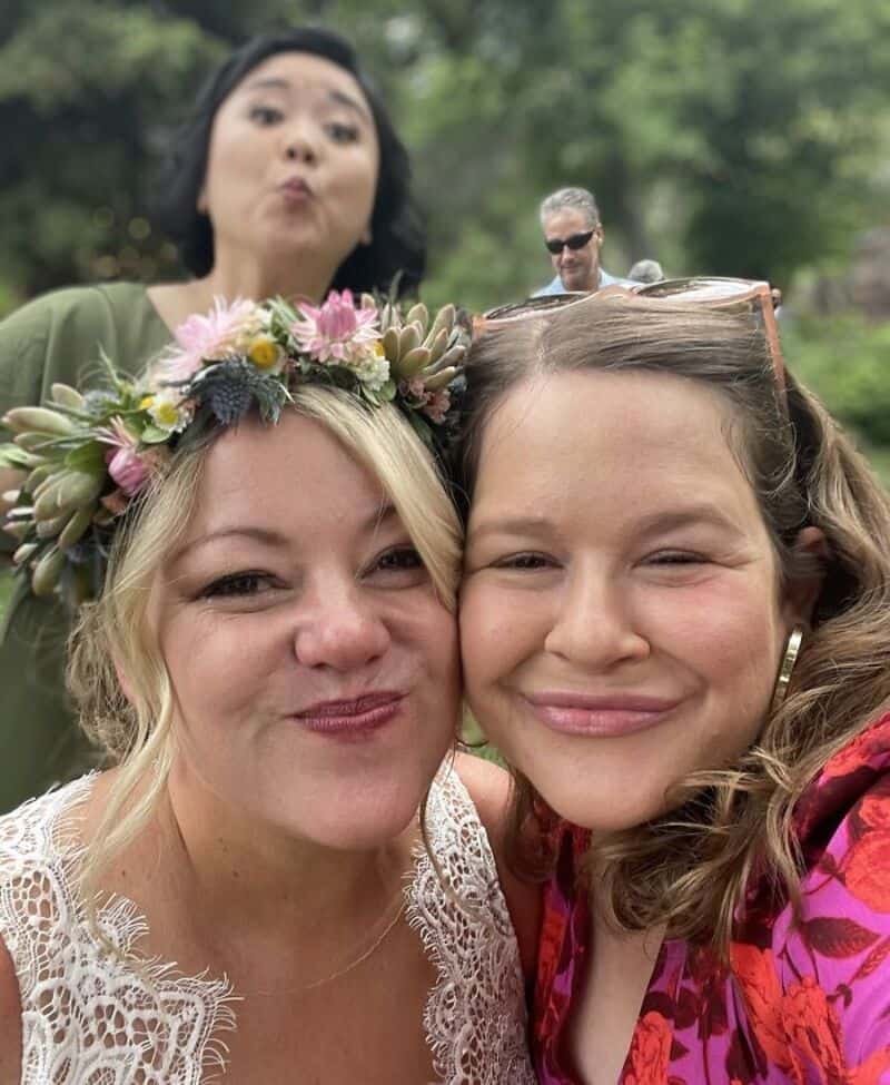 Two women smiling, one with a flower crown, and a woman making a funny face in the background.