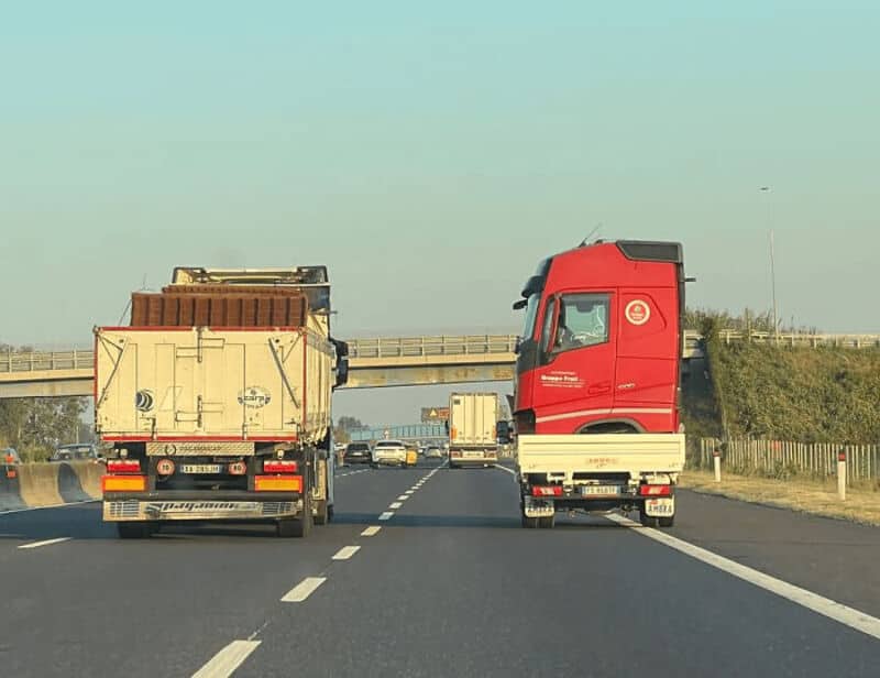 Two trucks on a highway, one red and one beige, with an overpass in the background.