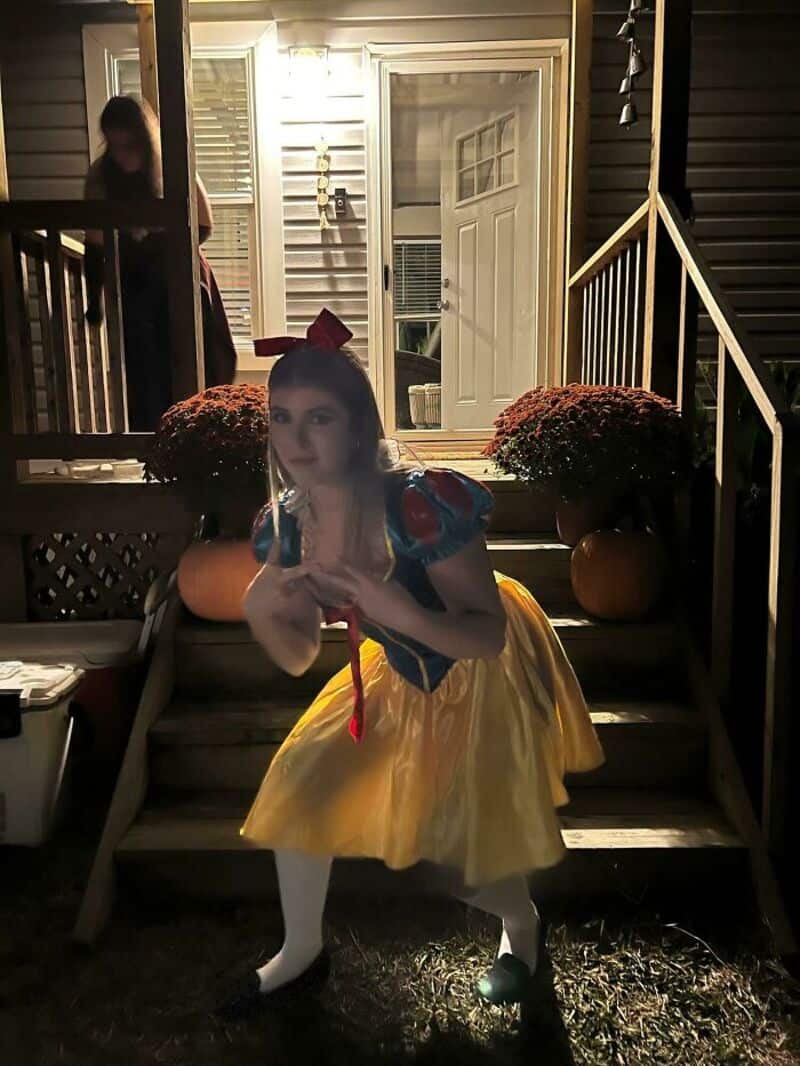 Girl dressed as Snow White on porch at night with potted plants and open door behind.