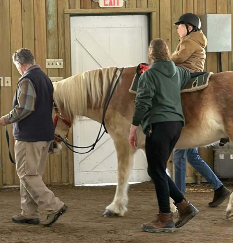 Child riding a horse with adults guiding inside a wooden stable.