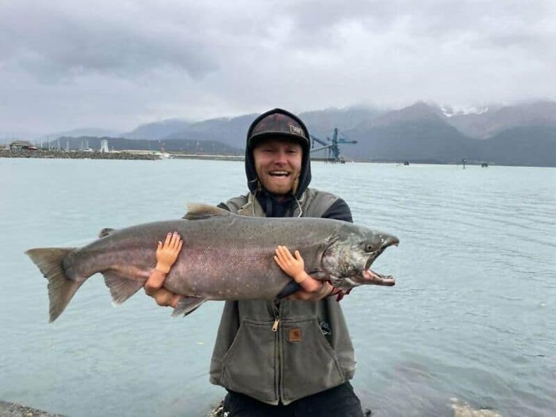 Man holding a large fish near the water with mountains in the background.