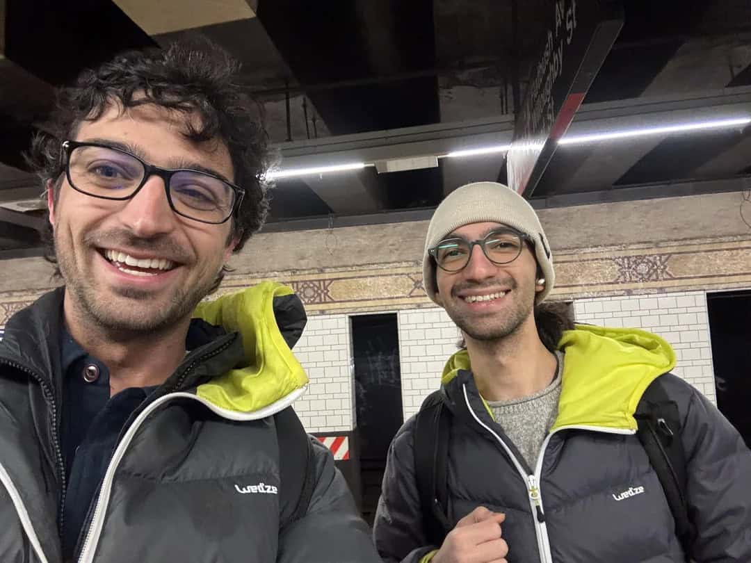 Two smiling men in jackets and glasses taking a selfie in a subway station.