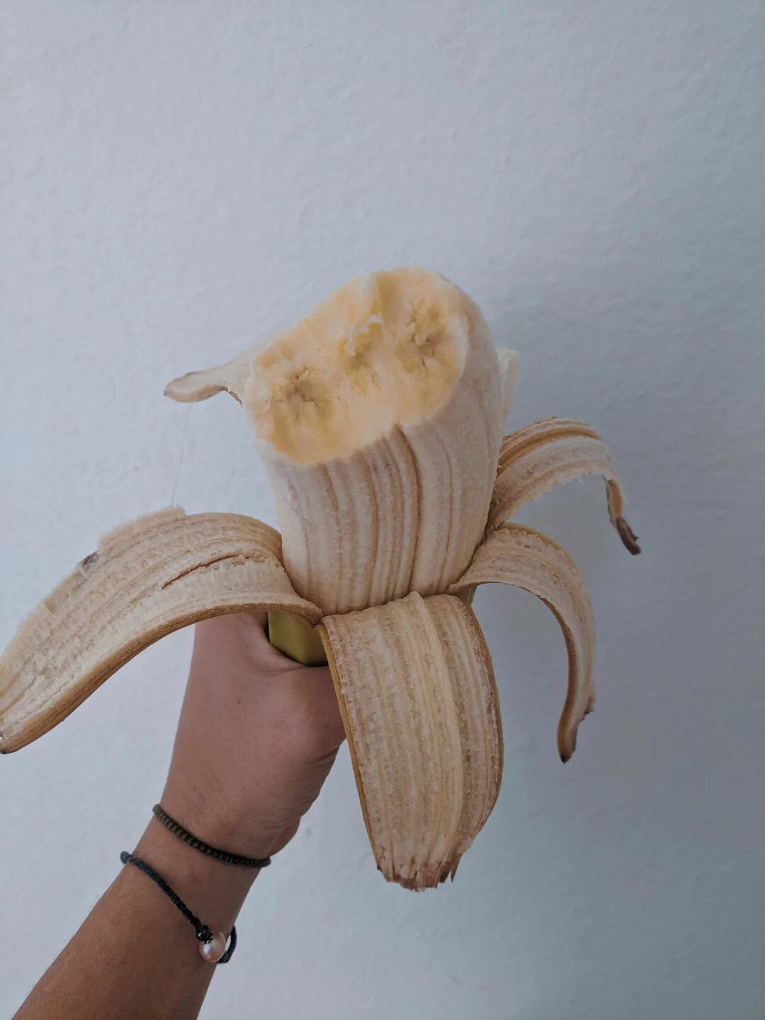 Banana peeled to reveal the inside, held by a hand against a plain wall.