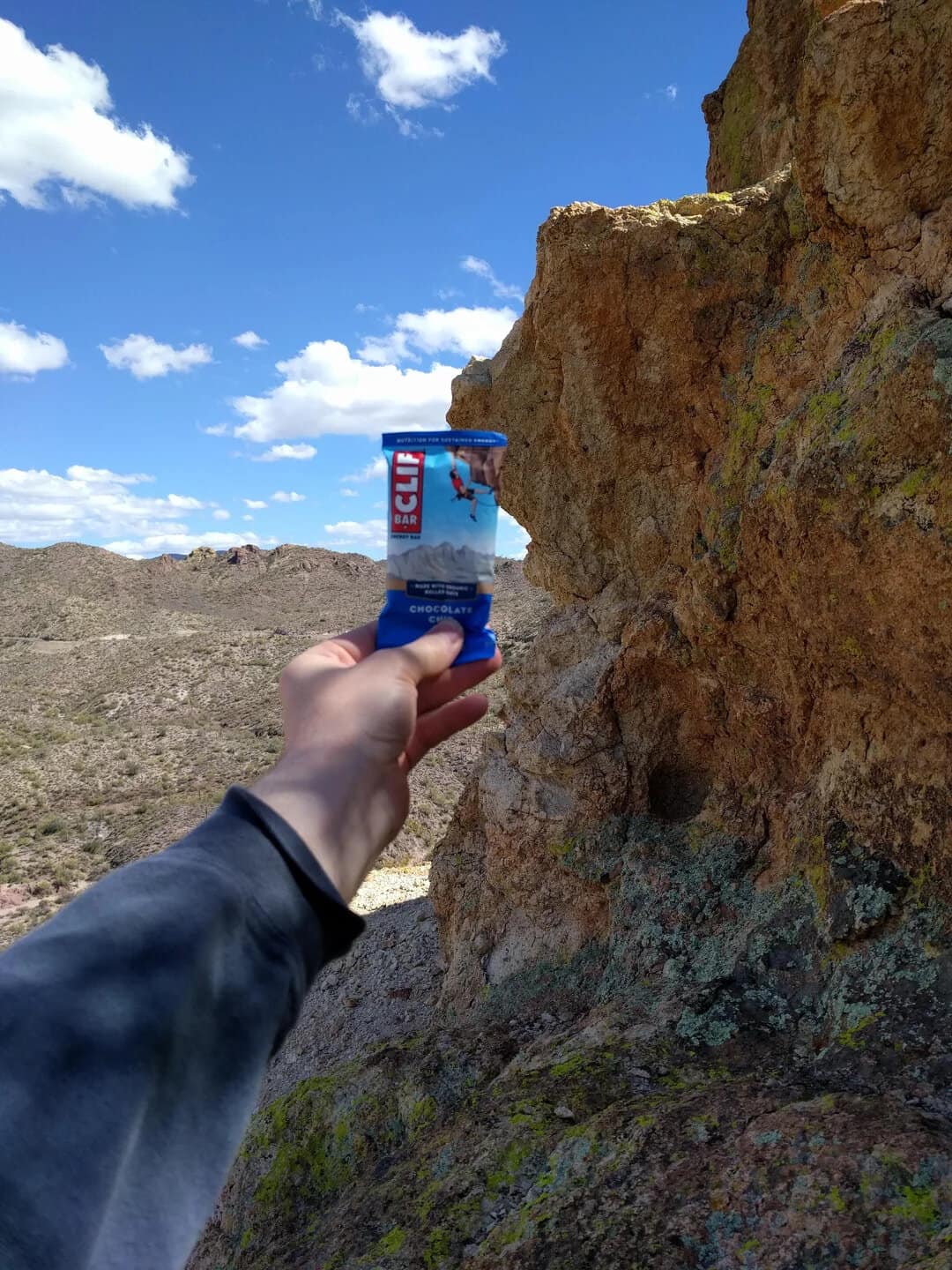 Person holding a Clif Bar in front of a rocky landscape and blue sky.