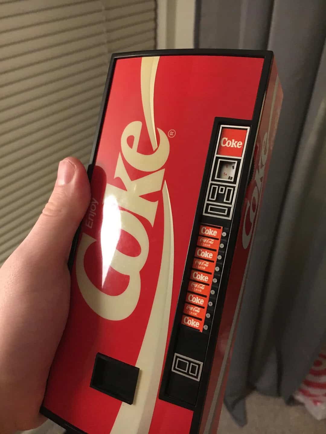 Hand holding a vintage Coca-Cola vending machine with a coin slot and buttons.