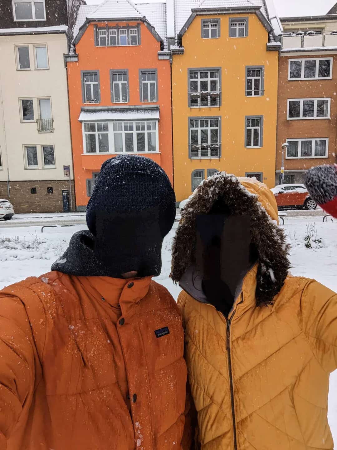Two people in winter jackets taking a selfie in front of colorful buildings during snow.
