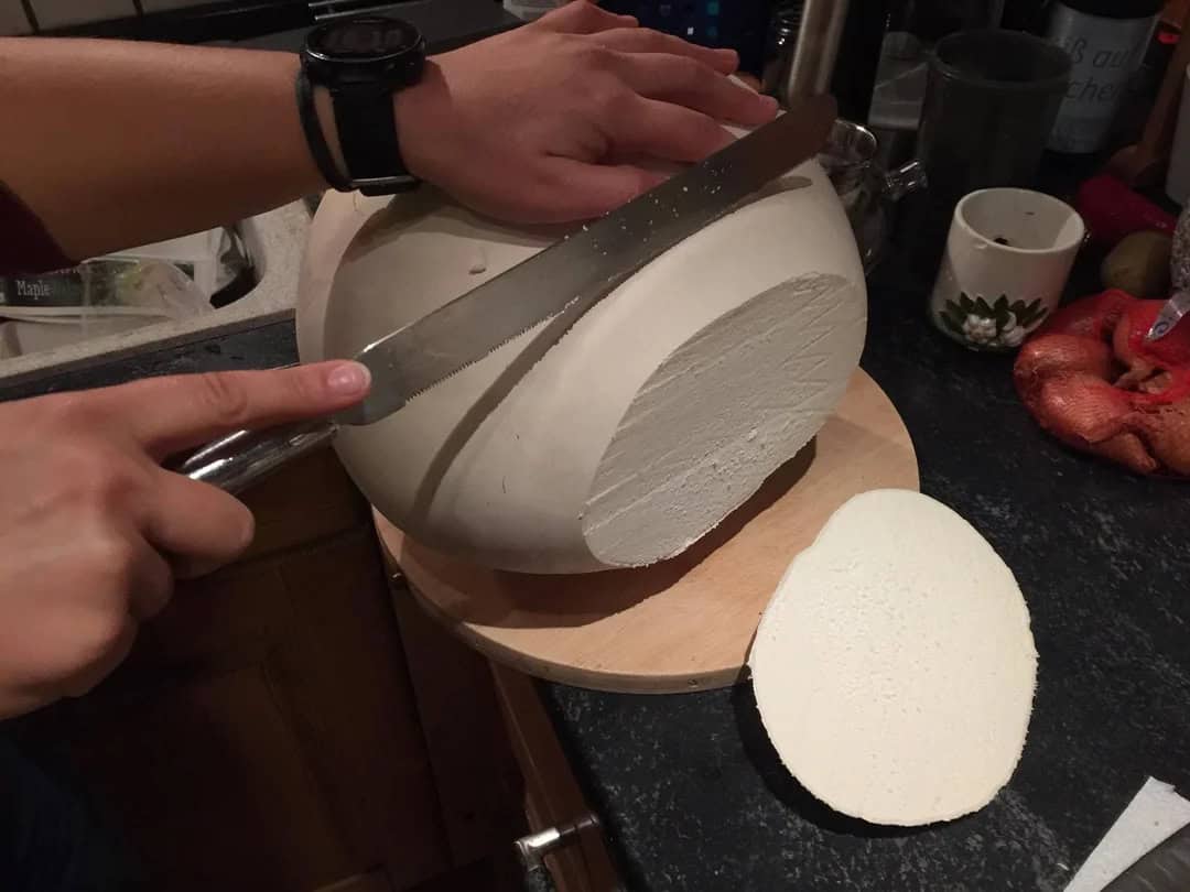 Person slicing a large block of white cheese on a wooden board in a kitchen.