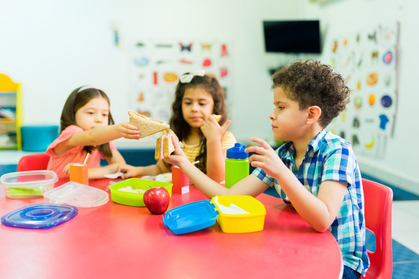 Kids sharing a sandwich at a colorful table in a classroom.