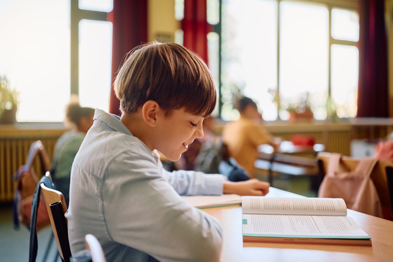 Young boy reading a book in a classroom with sunlight through windows.
