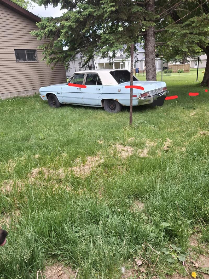 Vintage light blue car with red accents parked on grass near trees and houses.