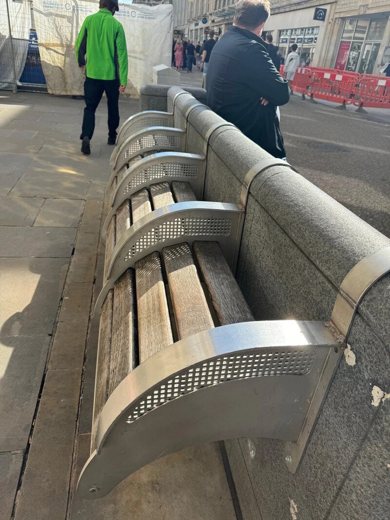 Public park bench with metal armrests and wooden slats on city sidewalk.