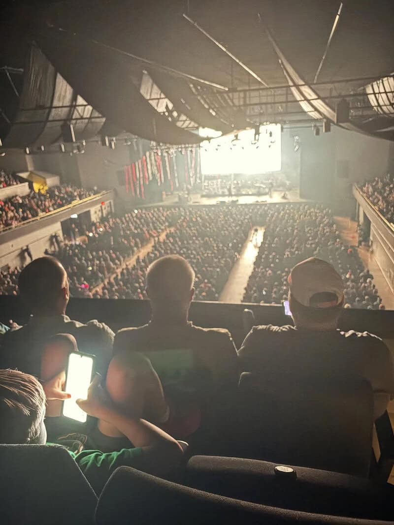 Large concert audience in a dark indoor arena with stage and bright screen.