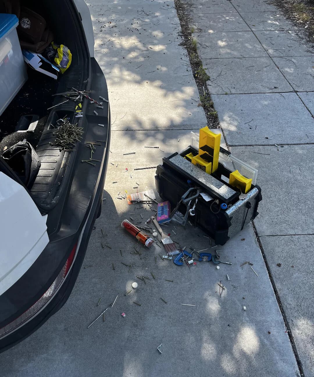 Tools and trash spilled on sidewalk next to open car trunk, with a toolbox and scattered items.