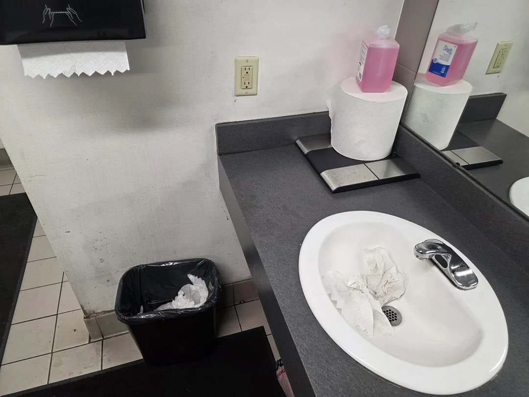 Dirty sink with crumpled paper and soap bottles, trash can nearby, in a public restroom.