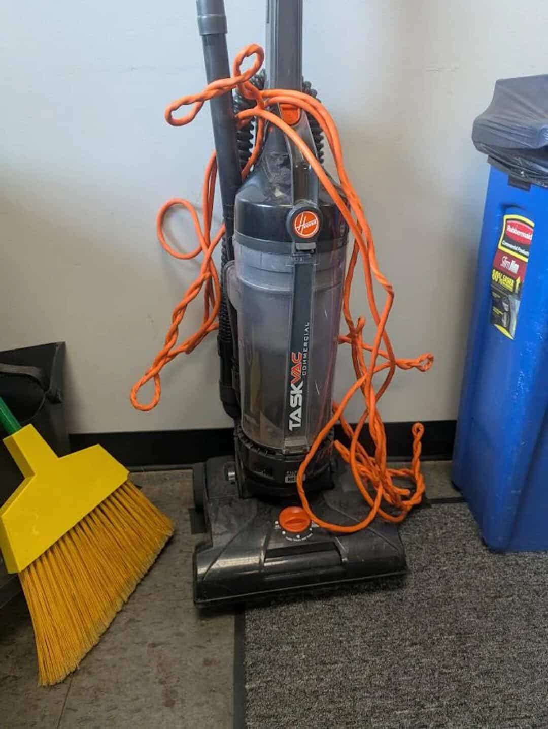 Vacuum cleaner with tangled orange cords next to a yellow broom and blue trash bin.