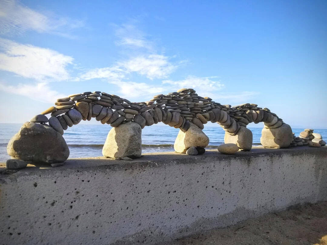 Stone arch bridge made of stacked stones on a beach under a blue sky.