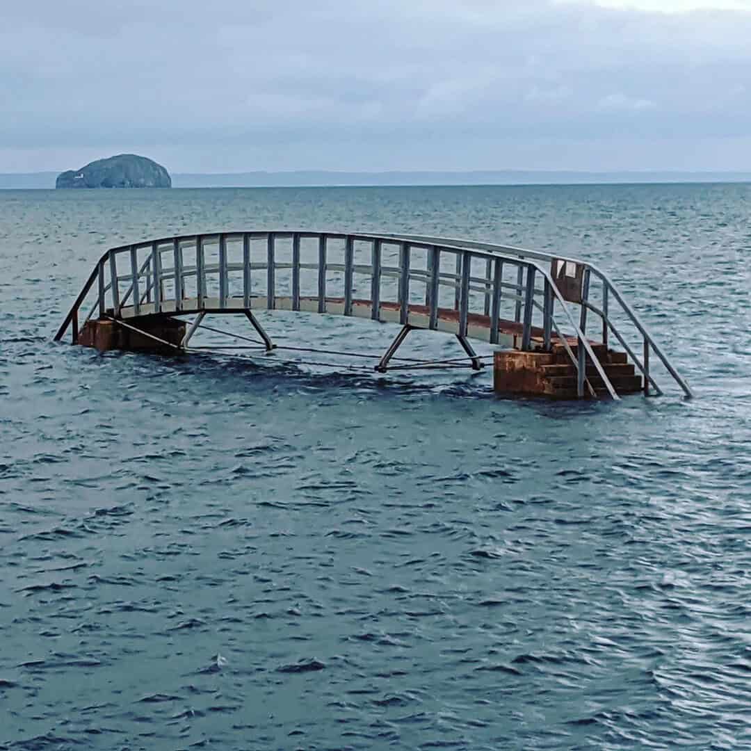 Submerged metal staircase in the ocean with a distant island and cloudy sky.