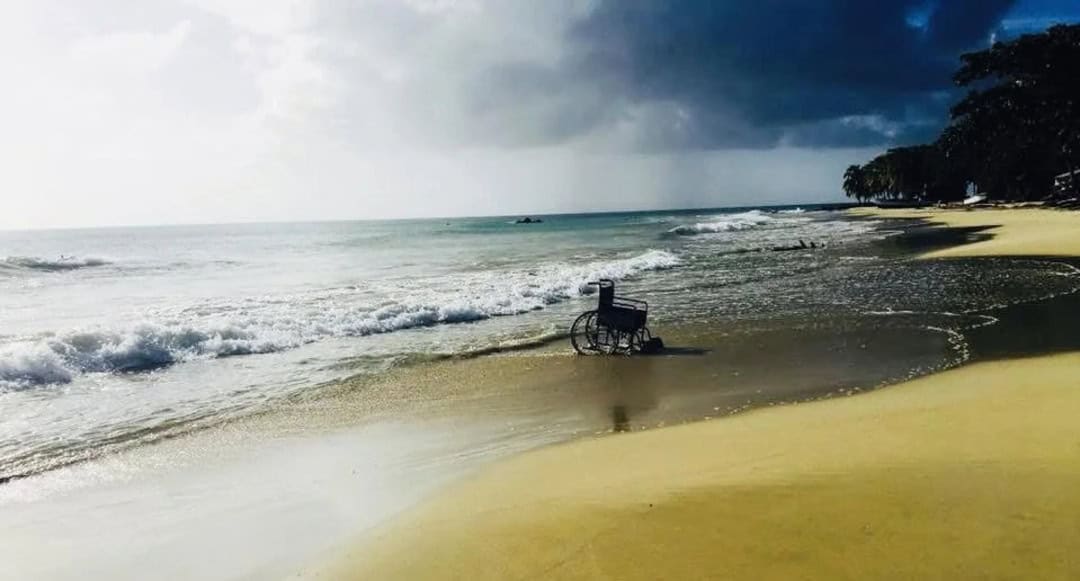 Empty wheelchair on a beach with dark storm clouds overhead and trees in the background.
