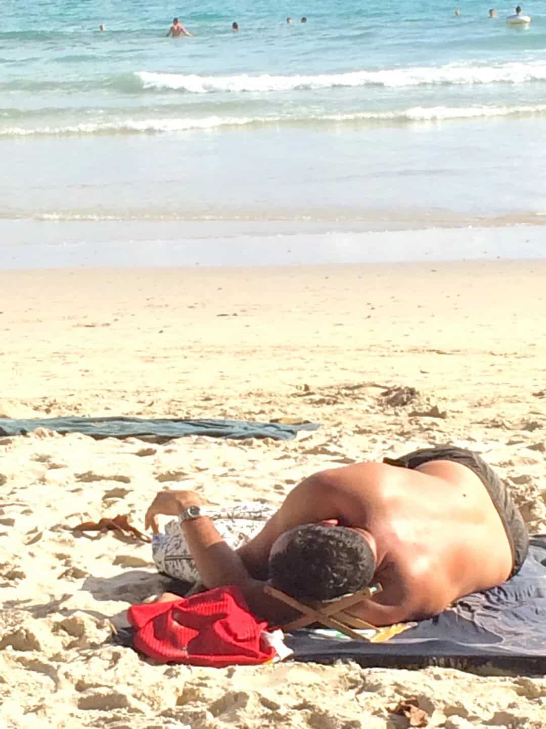 Person lying on the beach with a red bag, facing the ocean, in a relaxed pose.
